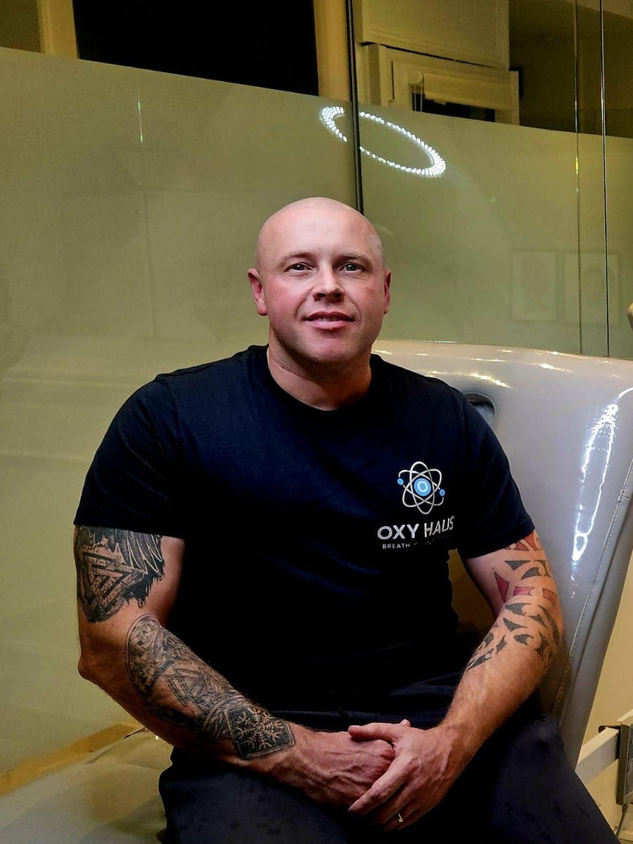 Stuart, founder of Oxy Haus, seated beside the hyperbaric chamber in an Oxy Haus shirt